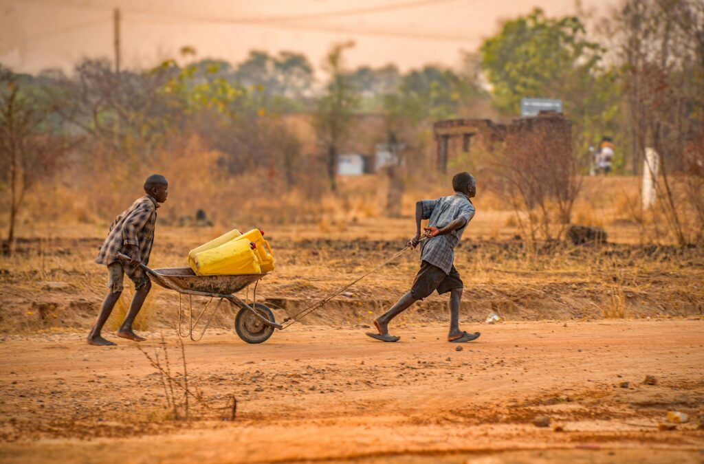 Two children transporting water containers on a wheelbarrow in rural Adjumani, Uganda.